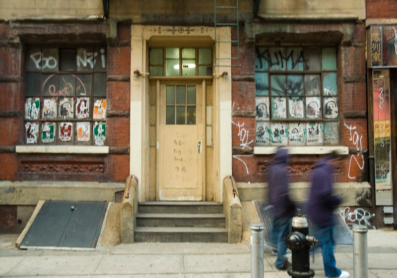 Two people walk past a building covered with graffiti and wheat paste posters.