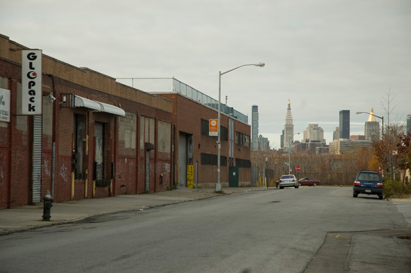 Two cars on an otherwise empty industrial street, with tall Manhattan buildings at back.