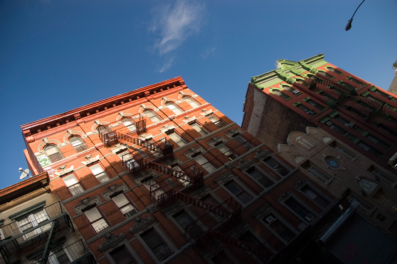 Rowhouses peek from shadows, in front of a clear blue sky.