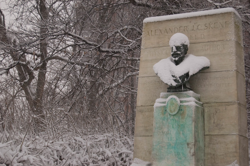 Snow coveres a bust on a pedestal, as well as surrounding trees.