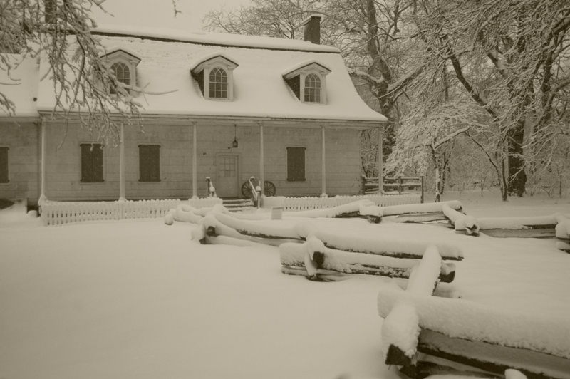 Snow covers an old wood frame house, its yard, and log walkway.