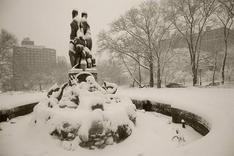 Snow covers the statues of a fountain in a plaza.
