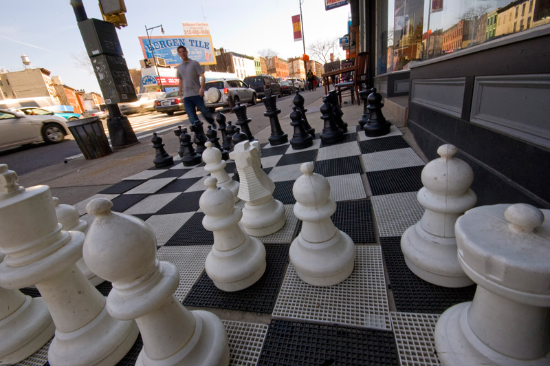 A plastic chess set with two foot tall pieces, in front of a store.