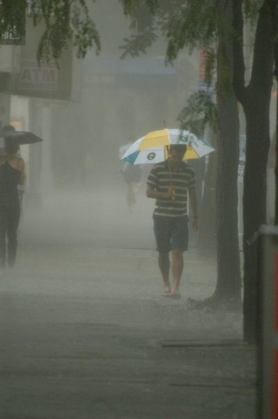 A man in shorts and sandals walks in a downpour, protected by his umbrella.