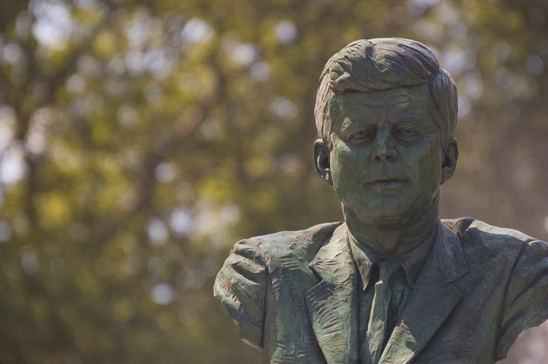 A bust of President John Kennedy, amidst trees.