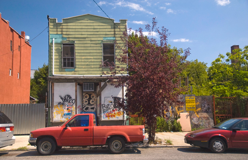 A red pick-up truck, parked in front of an abandoned, graffiti'd house.