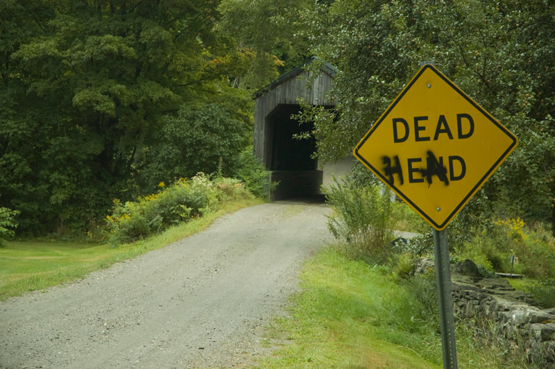 A 'Dead End' sign by a covered bridge, spray-painted to read 'Dead Head.'
