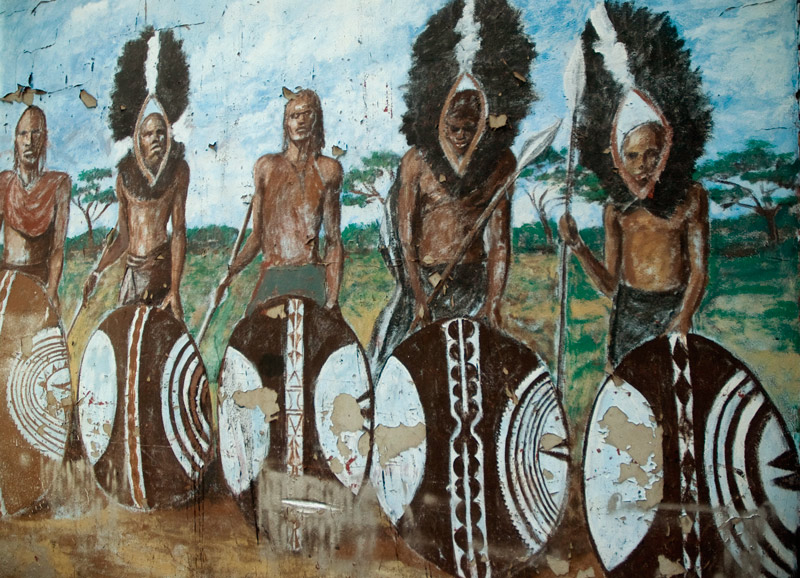 A mural with peeling paint shows African tribesmen with headdresses, spears, and shields.