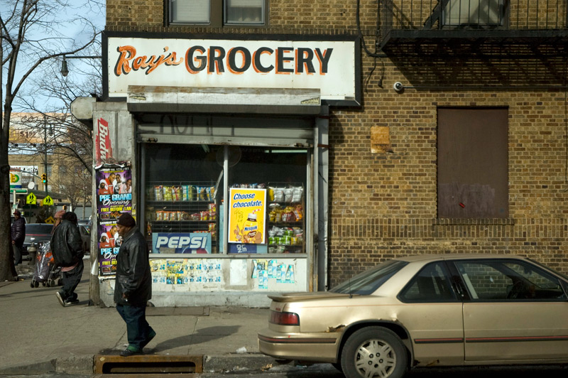 A man passes a bodega with a hand-lettered sign for Ray's Grocery.