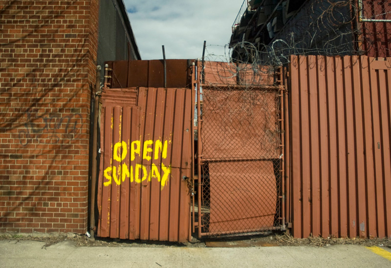A gate on a corrugated metal fence, with the hand-painted words 'Open Sunday.'