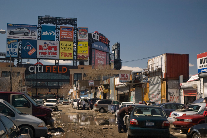 A street of mud and puddles, with auto shops and Citi Field.