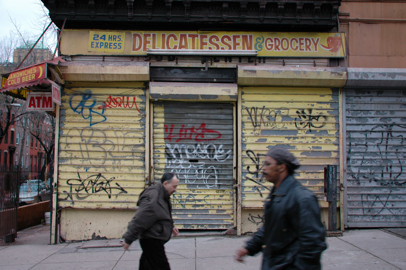 People walk past a closed bodega, with graffiti on its door front.