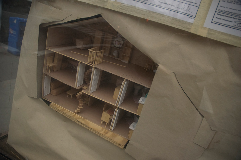 A doll house diorama peeks through brown paper in a storefront window.