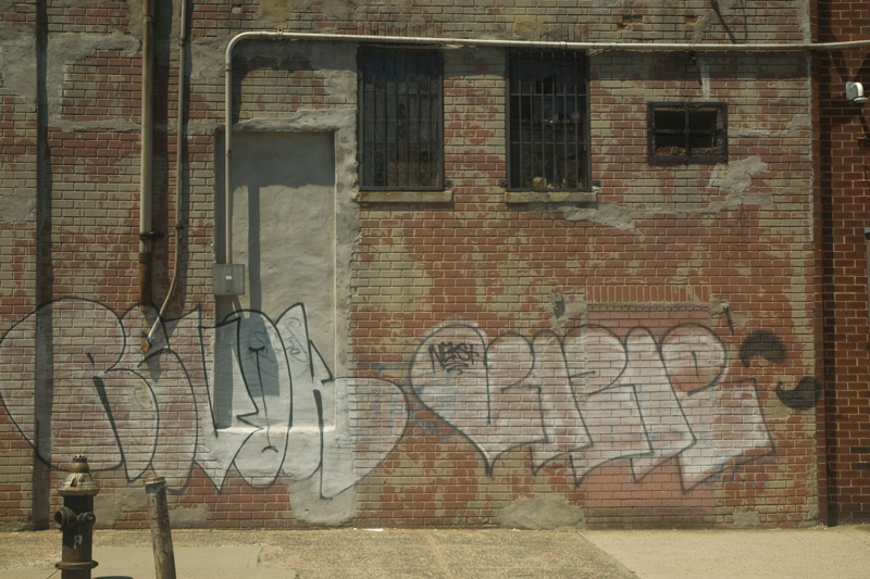 A brick wall, with piping and graffiti tags.