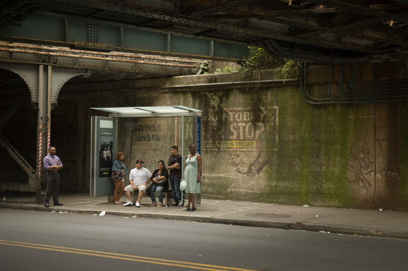 Six people at a bus stop, in front of a faded wall sign.