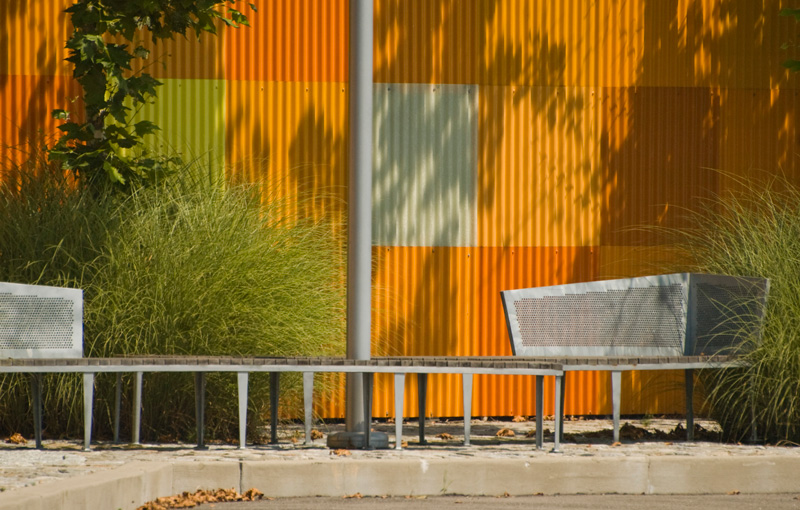 A metal bench in front of an orange corrugated metal wall.