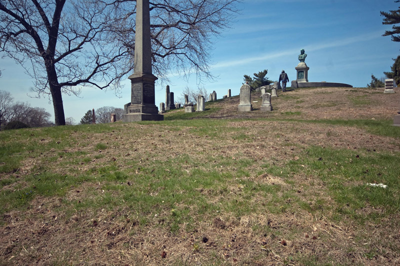 A man climbs a graveyard hill to a bust of Horace Greeley.