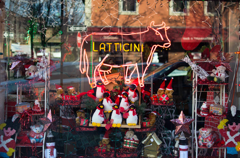 A neon sign in a delicatessen window reads 'Latticini.' There are Christmas items in the window.