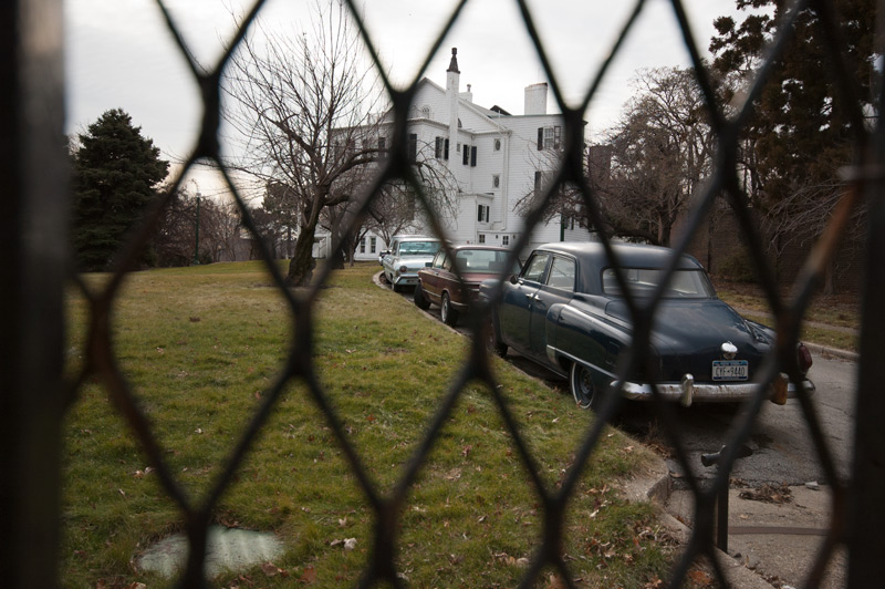 A white mansion behind a fence, with antique cars in the driveway.
