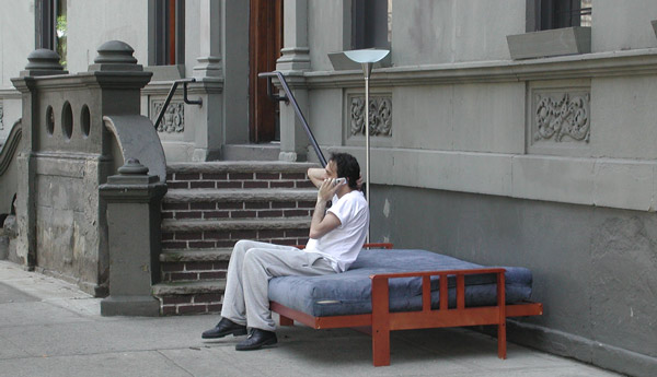 A man talks on
a cell phone while sitting on a bed, outdoors.