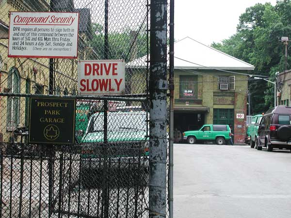 A chain link fence gate partly open, with trucks
and a garage building showing exposed brick.