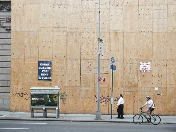 A man is dwarfed by a huge building which
is completely covered in a plywood front while it's under
renovation.
