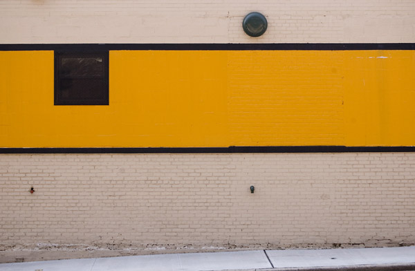 A beige, brick wall has a broad yellow stripe and a pipe or
so.